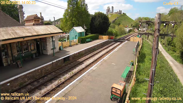 A small railway station platform with a stone building on the left, featuring a corrugated roof and windows. There are green benches and a yellow fence along the edge of the platform. Railway tracks run parallel to the platform, and a wooden utility pole with wires is on the right side. In the background, there is a grassy hill with ruins of an old castle at the top, surrounded by green trees under a partly cloudy sky.