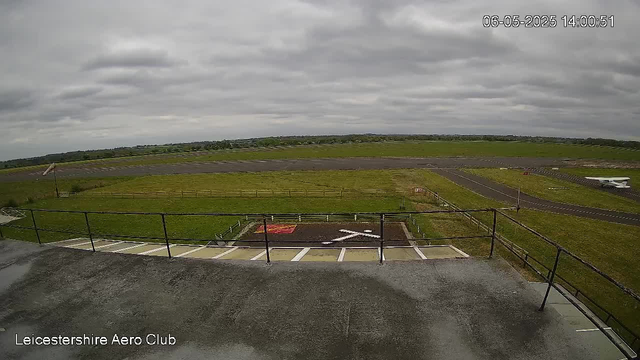 A view from a webcam showing an airfield with a runway and grassy areas under a cloudy sky. In the foreground, there is a small parking area with marked spaces and a red and white painted arrow indicating direction. To the right, a small aircraft is parked near the edge of the airfield. The scene is overcast with gray clouds, and the date and time are displayed in the upper right corner as June 5th, 2025, at 14:00:51. The bottom left corner has the text "Leicestershire Aero Club."