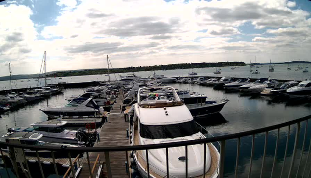 A marina filled with multiple boats and yachts docked along wooden piers. The water is calm and reflects the boats. The sky is partly cloudy with patches of blue visible. In the foreground, a railing curves along the edge of the dock.