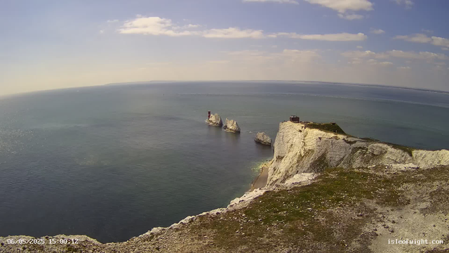 A scenic view of white chalk cliffs with patches of green grass at the top extends along a calm ocean. In the distance, three large rock formations rise from the water near the base of the cliffs, one of which has a small red and white lighthouse. The sky is partly cloudy with some rays of sunlight, and the horizon shows a long stretch of ocean meeting the sky. The photo is timestamped "06/05/2025 15:00:12" and includes a website "isleofight.com" in the bottom corner.