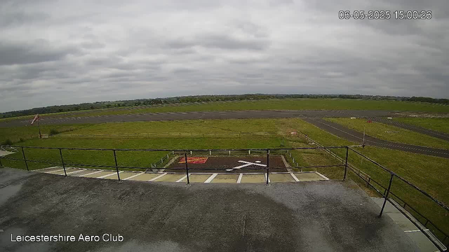 A view from a webcam showing an empty outdoor area at LeicesterShire Aero Club. In the foreground, there is a flat concrete surface with a metal railing and a pathway with an arrow painted on the ground indicating a left turn. Beyond the railing, a grassy field extends toward a small, paved tarmac area with a windsock on a tall pole. In the background, there are runways and taxiways stretching across the landscape under a cloudy sky. The timestamp in the top right corner indicates the date and time as June 5, 2025, at 15:00:26.