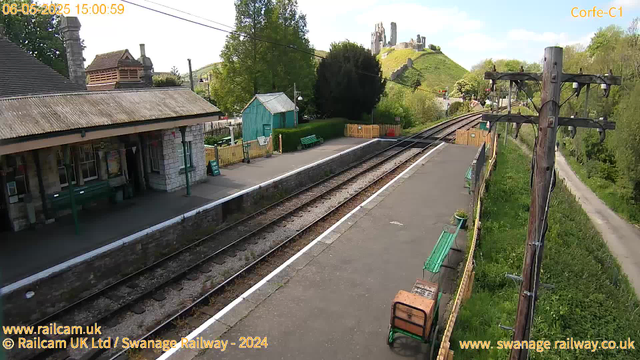 A small railway station platform with a building on the left side, featuring a stone wall and a roof. There are benches and a small teal-colored shed behind a hedge. The railway tracks run parallel to the platform, with a wooden fence and some benches along the edge. On the right side, a large wooden utility pole with electrical wires is next to a grassy area. In the background, a hill with ruins or ancient structures sits under a partly cloudy sky with some scattered clouds. The date and time displayed are May 6, 2025, at 15:00:59.