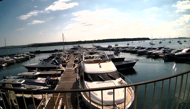 A marina filled with numerous boats and yachts docked along a wooden pier, with calm water reflecting the vessels. In the background, there is a body of water extending toward the horizon under a partly cloudy sky. The photo is taken from a vantage point behind a curved railing.