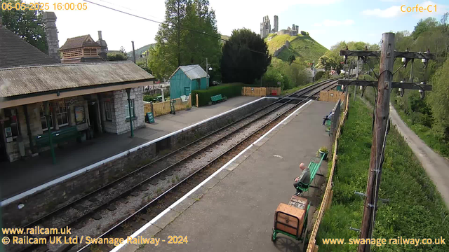 A small train station platform with a stone building on the left, featuring a sloped roof and large windows. There are a few benches, one occupied by a person wearing a gray jacket, sitting and looking at their phone. Along the platform's edge, a white line marks the boundary. To the right, wooden utility poles with electrical wires run along a narrow grassy path bordered by trees and bushes. In the background, a green hilltop features ruins of an old castle or similar structure, with a blue sky overhead. The date and time are displayed at the top left as "06-05-2025 16:00:05", and labels "Corfe-C1" and website information are at the top right and bottom left.