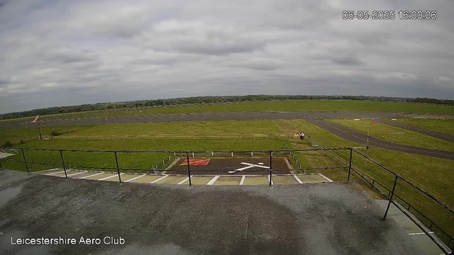 This is a wide view of a grassy airfield under a cloudy sky, captured from a webcam. In the foreground, there is a concrete observation deck with a black railing, and a helicopter landing pad marked with a white 'H' and a red marking nearby. To the right of the pad, there are a few small vehicles or equipment. Further out, there is a taxiway with painted markings, and grassy runs extending into the distance. A small windsock on a pole is to the left, indicating wind direction. The image is labeled "Leicestershire Aero Club" in the lower left corner, and the timestamp in the upper right corner shows it was taken on June 5, 2025, at 4:00 PM.
