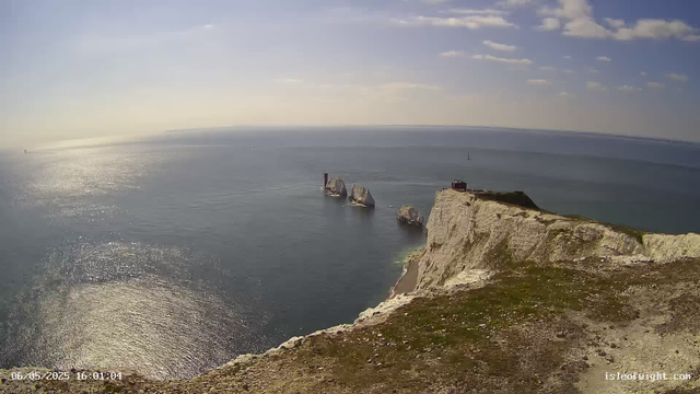 A view of white chalk cliffs with patches of green grass on top, rising above the sea. The cliffs extend into the water with a few large rocks or sea stacks visible just offshore. The sea is calm with gentle ripples, and the sunlight reflects off its surface, creating a shimmering effect. The sky is mostly clear with a few scattered clouds. The image is timestamped "06/05/2025 16:01:04" and has text in the bottom right corner that reads "isleofwight.com".