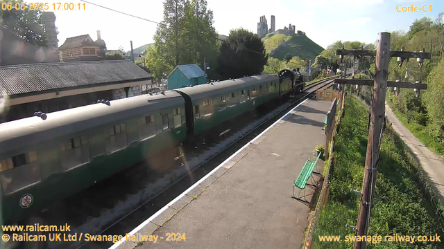 A green-colored train with multiple carriages is stopped at a small rural train station. The platform is narrow with a green bench and a potted plant. To the right, there are wooden electrical poles with multiple wires. In the background, there is a hill with standing stone ruins at the top, surrounded by greenery and trees. The sky is clear and sunny. Some buildings with metal roofs and a small blue shed are visible behind the train. The timestamp on the image indicates the date and time as June 5, 2025, at 17:00:11.