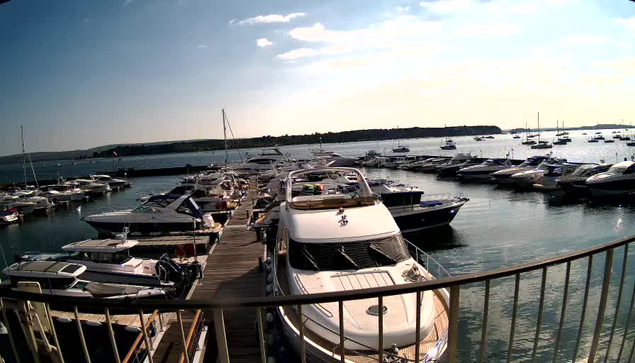 A marina with multiple boats and yachts docked along wooden piers extending into calm water, with an expansive sky and a distant shoreline in the background.