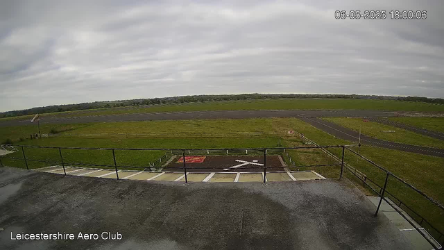 An outdoor view from a webcam at Leicestershire Aero Club shows a vast open field with a runway extending into the distance under a cloudy sky. In the foreground, there is a small platform with a railing, and a painted helipad marking on the ground. The timestamp indicates the date and time as June 5, 2025, at 6:00 PM.
