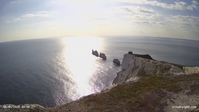 A scenic view of white chalk cliffs overlooking the ocean. The sun is low in the sky, reflecting brightly on the water. A small tower is visible on one of the rocky outcrops extending into the sea. The overall atmosphere is calm and peaceful with partly cloudy skies.