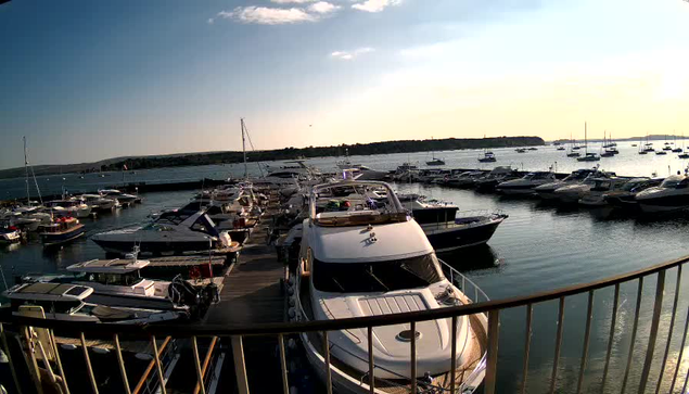 A marina filled with numerous boats and yachts docked along wooden piers, with calm water reflecting the vessels. In the background, an island or shoreline extends into the horizon under a sky with a warm, setting sun. A curved railing is visible in the foreground, suggesting the photo was taken from a balcony or deck.