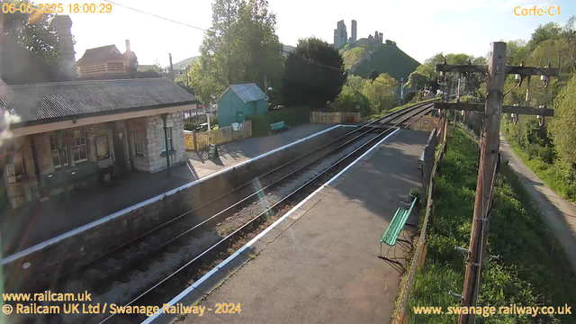 A small train station platform is visible, with a building on the left featuring a stone exterior and a corrugated metal roof. There is a green bench on the platform, and behind it, a small turquoise shed. The platform is bordered by a white line along the edge of the track. Multiple railway tracks run parallel, curving slightly to the right in the distance, with wooden utility poles and overhead power lines along the right side of the tracks. In the background, on a hill, ancient ruins or castle structures are partially visible amidst green trees, and the sky is clear and bright. The image is timestamped 18:00:29 on June 5, 2025.