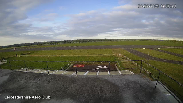 A wide view of an open grassy airfield with a parking area in the foreground, marked with an 'X' to designate a helipad. There is a small painted sign on the grass, and a paved runway extending into the distance. The sky is partly cloudy with patches of blue, and the surrounding landscape is flat with distant trees on the horizon. A timestamp in the top right corner indicates the date and time as June 5, 2025, at 19:00:23. The bottom left corner reads "Leicestershire Aero Club".
