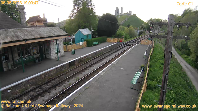 An empty train station platform with a building on the left, a small turquoise shed, greenery, and a castle ruin on a hill in the background. There are benches, a wooden fence, and a tall wooden telegraph pole with wires on the right. The sky is clear, and the scene appears to be during late afternoon or early evening.