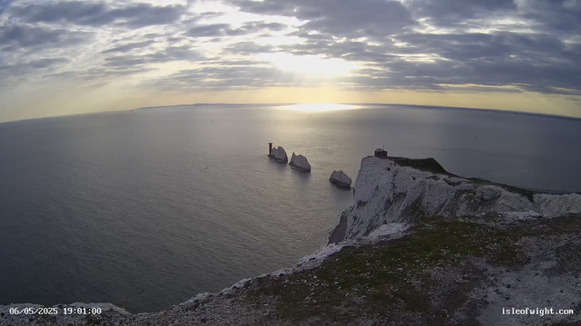 A view from a webcam shows white chalk cliffs on the right, with a building or lookout on top. The cliffs drop sharply into the calm sea below. In the distance, there are several large sea stacks rising from the water. The sky is partly cloudy with the sun shining through the clouds, creating a reflection on the water. The timestamp indicates the date and time as June 5th, 2025, at 7:01 PM.