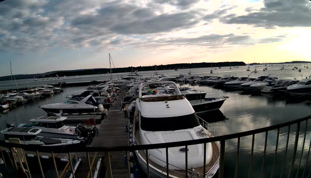 A marina filled with numerous boats docked along wooden piers under a cloudy sky, with the water reflecting the dark clouds and the few patches of light from the setting or rising sun in the distance.