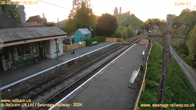 A railway station platform at sunset, with a small stone building on the left, a green bench, and a fenced garden area behind it. Train tracks run parallel to the platform, stretching into the distance. To the right, wooden telegraph poles with insulators and wires run alongside a narrow path surrounded by greenery. In the background, a hill with a castle or ruins sits atop, illuminated by the setting sun. The overall scene is calm and peaceful.
