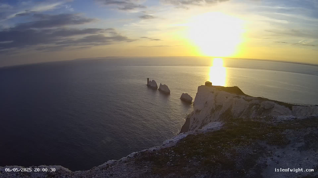 A panoramic view of a coastal landscape during sunset, showing white chalk cliffs extending into the sea. The sun is low on the horizon, casting a golden reflection on the water. Rock formations, including a prominent sea stack with a lighthouse, rise from the ocean near the base of the cliffs. The sky has scattered clouds with shades of blue, gray, and orange. The timestamp at the bottom left reads "06/05/2025 20:00:30," and the website "isleofwight.com" is displayed at the bottom right.