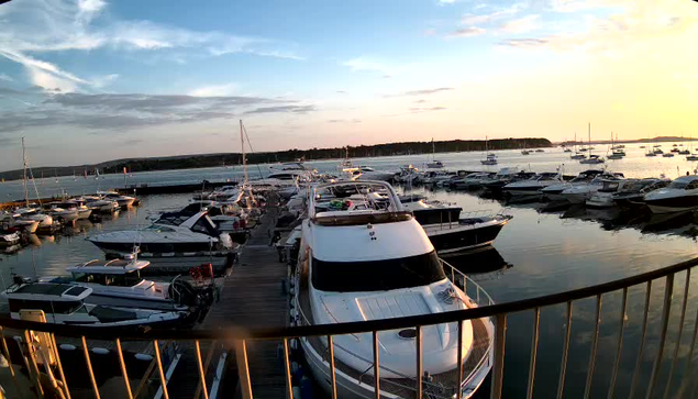 A marina filled with various boats and yachts docked at the piers during sunset. The sky is partly cloudy with the sun low on the horizon, casting a warm glow over the scene. The calm water reflects the boats and the sky. A railing is visible in the foreground.