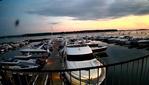 A marina filled with various boats and yachts docked along wooden piers, with calm water reflecting the boats. The sky shows a mix of dark clouds and a soft glow from the setting or rising sun, creating a serene atmosphere. A black metal railing is visible in the foreground.
