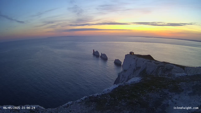 A view of white chalk cliffs overlooking the ocean at sunset, with a small lighthouse on top of the cliffs and several sea stacks standing in the water near the coast. The sky is partly cloudy with hues of orange, yellow, and blue. The date and time are displayed in the bottom left corner: 06/05/2025 21:00:24. The website "isleofwight.com" is visible in the bottom right corner.