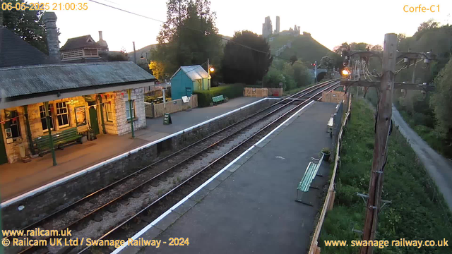 A small train station platform is visible in the foreground with a stone building on the left side that has a green door and windows, and a green bench outside. There are two green benches along the platform, one near the building and another further down. A few potted plants are placed next to the benches. A "WAY OUT" sign is near the building entrance. The train tracks run through the middle of the image from bottom to top, with a fence running alongside the tracks on the right side. A row of wooden benches and a large wooden utility pole with electrical wires is on the right side of the platform. In the background, the sky is bright, and there are trees, a small turquoise-colored shed, and a hill with what appears to be a castle or ruins on top. The timestamp indicates the photo was taken on June 5, 2025, at 21:00.