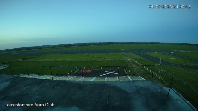 An outdoor view from a webcam showing a grassy field and a small runway or helipad with a white helicopter symbol. There is a black railing in the foreground and a wide open landscape extending into the distance under a clear sky. The timestamp indicates the date and time as June 5, 2025, at 9:00 PM, and the bottom left corner contains the text "Leicestershire Aero Club."