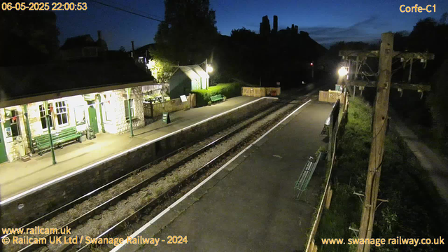 A quiet train station platform at night with two benches, a small building with windows and a door, illuminated by bright lights. The railway tracks run through the center, and a tall wooden pole with electrical wires on the right side. There are a few trees and houses silhouetted against the darkening sky in the background. The timestamp shows it is 22:00 on May 6, 2025.