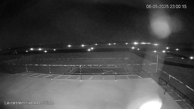 A nighttime view of an outdoor sports field at the Leicestershire Aero Club, viewed from a webcam. The field is empty, with fencing around it, and parking spaces are visible in the foreground. Several bright lights are scattered across the background, illuminating the dark sky, and the full moon is visible in the upper right corner. The date and time stamp in the upper right corner reads "06-05-2025 23:00:15."