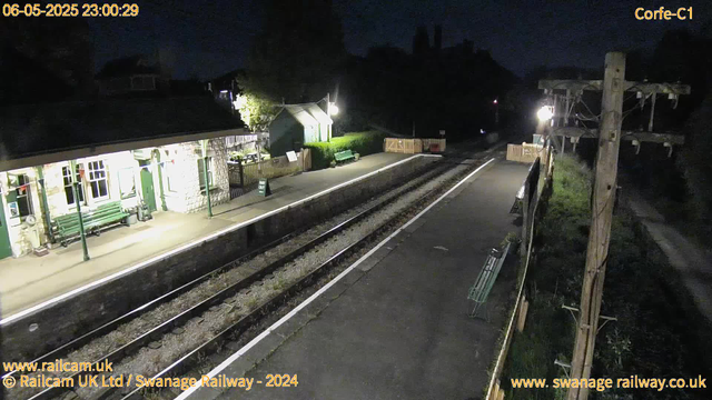 A nighttime scene at a small railway station. The platform is illuminated by a few bright lights, with empty benches and a small green building. A "Way Out" sign is visible on the platform. The railway tracks run parallel to the platform, with some fencing and utility poles along the side. The sky is dark, and the area appears quiet.