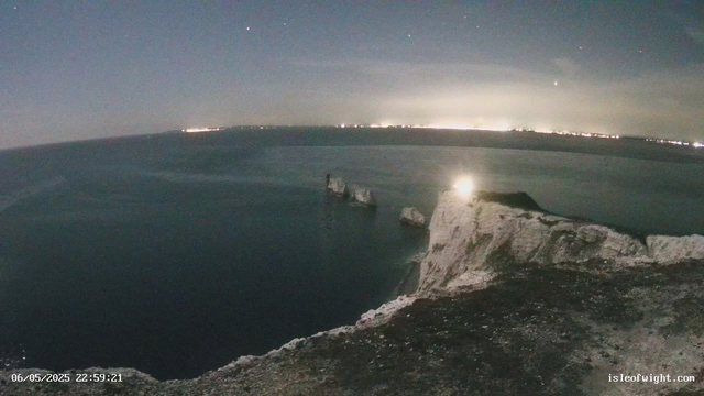 A nighttime view of a cliffside overlooking the ocean. There are white chalk cliffs with some rock formations protruding into the water. The sky is dark with a few visible stars, and the horizon is faintly illuminated by distant city lights or lighthouse glow. The scene appears calm with minimal wave activity. The timestamp at the bottom left indicates it was taken on June 5, 2025, at 10:59 PM.