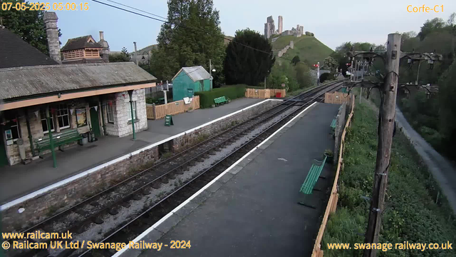 A small railway station platform with green benches, a stone building with a tiled roof, and a small green shed. There are safety barriers on the sides of the platform, a row of electrical poles with wires overhead, and a grassy hill with ruins of an old castle in the background. The scene appears to be during early morning.