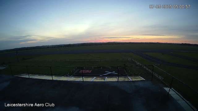 A wide view of an airfield during sunrise with a flat grassy landscape stretching to the horizon. In the foreground, there is a helipad marked with a white helicopter symbol, surrounded by a safety railing. The sky is partly cloudy with soft pastel colors. The date and time displayed in the top right corner are "07-05-2025 05:00:57," and the bottom left corner contains the text "Leicestershire Aero Club."