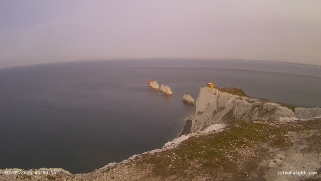 A view of the white chalk cliffs along the coastline with a small lighthouse on the edge of the cliffs. There are several large rocks in the water near the shore. The sky is mostly clear with a hint of haze near the horizon. The date and time stamp in the corner indicates "07/05/2025 05:00:56," and the website "isleofwight.com" is visible in the bottom right corner.