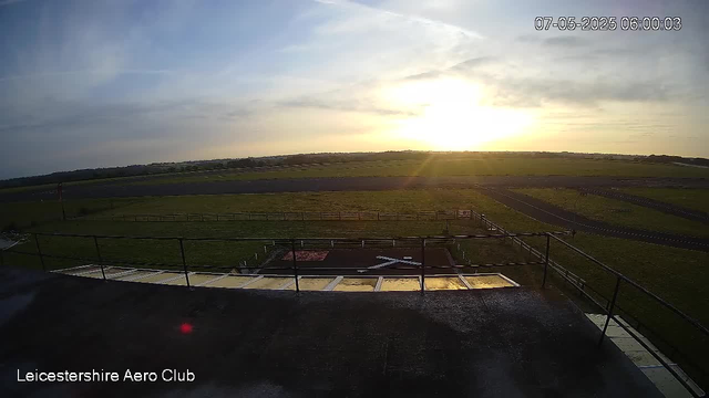A sunrise or sunset over a flat, open landscape with grassy fields and some paved areas. The sky has scattered clouds, and the sun is low on the horizon, casting a warm glow. There are railings and a small platform or deck in the foreground, with the text "Leicestershire Aero Club" in the bottom left corner. The timestamp in the top right corner indicates the date and time as July 5, 2025, at 6:00 AM.
