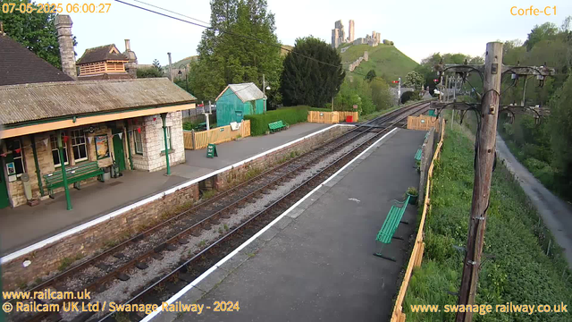 A small train station platform with a building on the left, featuring a green door and window frames. There are green benches along the platform, and a green "Way Out" sign near the middle. The railway tracks run through the center of the image, bordered by a low brick wall. On the right side, there is a tall wooden telegraph pole with electrical wires overhead. The surrounding area is lush with green trees and grass, and a hill with ancient ruins or castle remains is visible in the background under a clear sky. The camera timestamp shows "07-05-2025 06:00:27."