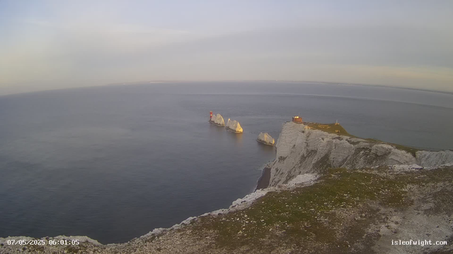 A view of white chalk cliffs along the coastline, with a grassy top and a small building on the edge. Several large white rock formations are located in the sea just offshore. The sky is cloudy and the sea appears calm. The image includes a timestamp of "07/05/2025 06:01:05" in the bottom left corner.