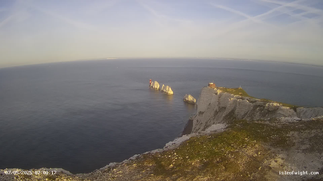 A panoramic view of white chalk cliffs rising from the calm ocean, with several large sea stacks just offshore. A small lighthouse or navigational aid is visible on the right side atop the cliffs, and the sky is clear with some wispy clouds. The photo appears to be taken early in the morning, as indicated by the timestamp.