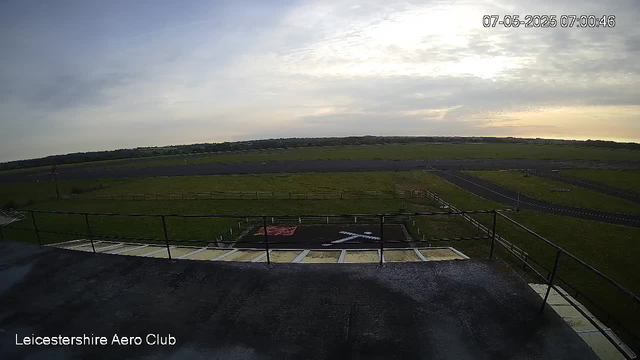 A wide view of an open grassy field with a small helicopter landing pad marked "H" in white. There are metal railings along the edge of a building or balcony in the foreground. The sky is mostly cloudy with some sunlight breaking through, and a rural landscape stretches into the distance. The timestamp in the upper right corner shows "07-05-2025 07:00:46," and the bottom left corner has the text "Leicestershire Aero Club."