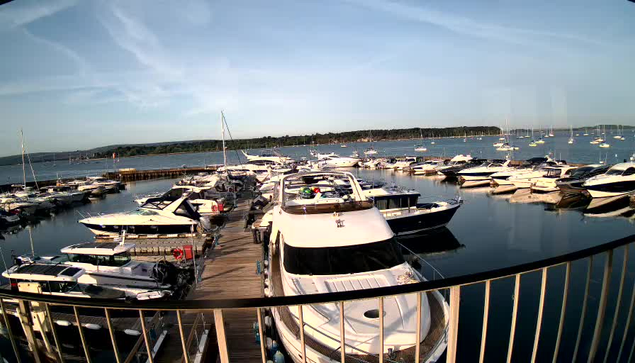 A marina filled with numerous boats and yachts docked along wooden piers. The water is calm and reflects the vessels. In the background, there is a distant tree-lined shoreline under a partly cloudy sky. A metal railing is visible at the bottom of the image, indicating the photo was taken from a boat or platform near the dock.