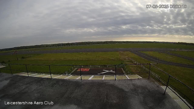 An outdoor view from a webcam at Leicestershire Aero Club showing a cloudy sky over an open grassy area with runways and taxiways in the distance. The foreground includes a landing pad with a helipad marked with a white helicopter symbol and an "X" across it, surrounded by metal railings. The timestamp indicates the date and time as July 5, 2025, at 8:00 AM.