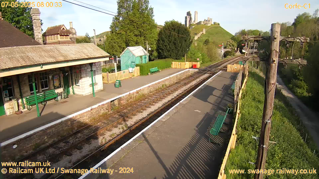A small train station with a platform, green benches, a green historic-style building, and a surrounding fence. Tracks run alongside the platform, and there are trees and a lush green hill with ruins of a castle on top in the background. A wooden fence and utility poles with wires are visible on the right side of the image. The scene is bathed in bright, early morning sunlight.