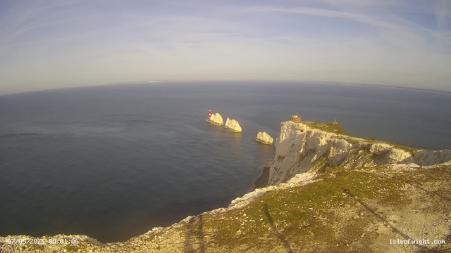 A panoramic view of white chalk cliffs overlooking a calm ocean. Several large white rock formations are visible in the water, with a red and white lighthouse on one of the rocks. A small building and a grassy area are situated atop the cliffs. The sky is mostly clear with a few wispy clouds.