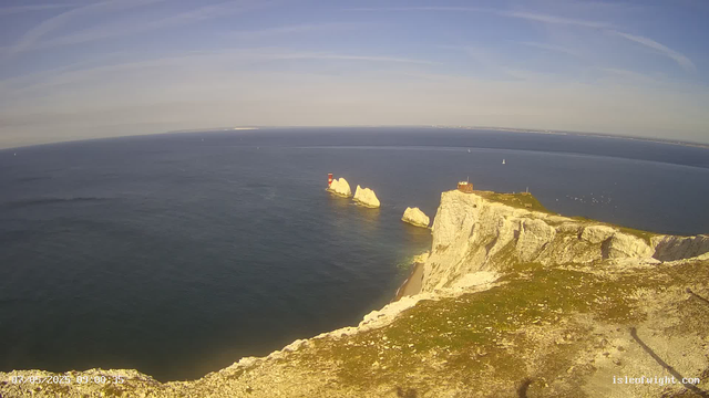 A wide view of a coastal landscape featuring white chalk cliffs with green grassy tops, a lighthouse on the edge of the cliffs, and several large sea stacks in the water below. The ocean extends to the horizon, with a clear sky above. There are a few sailboats visible in the distance. The time and date are displayed in the lower left corner.