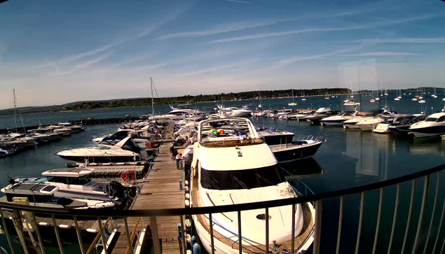 A marina filled with numerous boats and yachts docked along a wooden pier. The sky is clear with a few wispy clouds, and a distant shoreline is visible in the background. The water is calm and reflective.