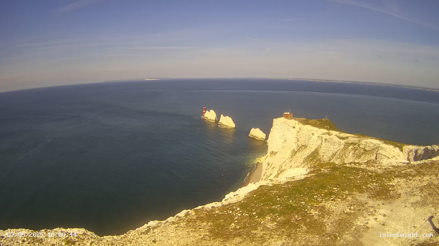 An aerial view of white chalk cliffs with green grass on top extend into the sea, with several large sea stacks rising from the water. A lighthouse with red and white stripes is located on one of the stacks. The sky is clear with a few wispy clouds, and the ocean stretches to the horizon.