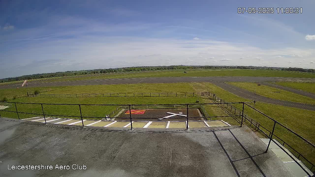 An empty airfield with a small parking area and safety railings in the foreground. The runway extends into the distance under a partly cloudy sky. The image is timestamped "07-05-2025 11:00:31" and labeled "Leicestershire Aero Club."