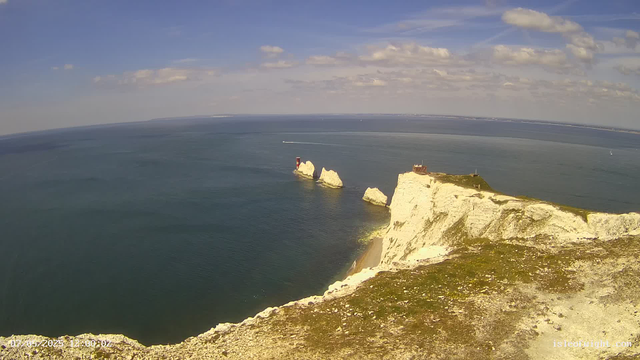 A scenic view of white chalk cliffs overlooking the ocean, with a small lighthouse on one of the cliffs and large rock formations rising from the water. The sky is partly cloudy with a few scattered clouds. In the distance, the horizon line separates the sea from the sky.