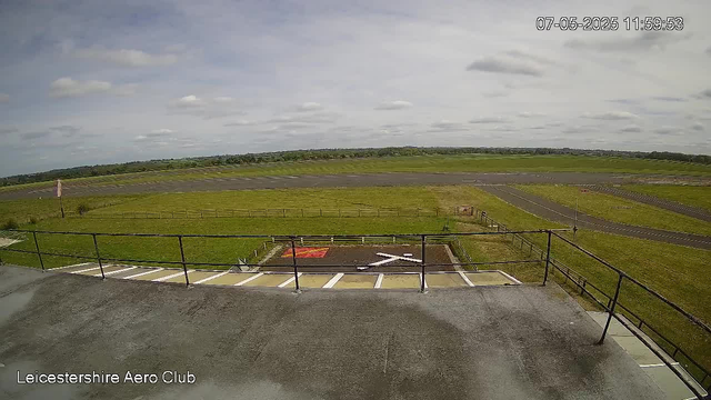 A wide outdoor view from a webcam shows a grassy field with a paved area and parking spots in the foreground, and a runway or taxiway extending into the distance under a partly cloudy sky. The image is labeled "Leicestershire Aero Club" in the bottom left corner, and a timestamp in the top right corner reads "07-05-2025 11:59:53."