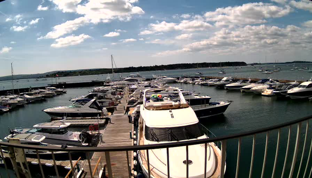 A marina filled with numerous boats and yachts docked along wooden piers, with calm water and a cloudy sky overhead. The image appears to be taken from a balcony or a viewing platform with a black railing in the foreground.